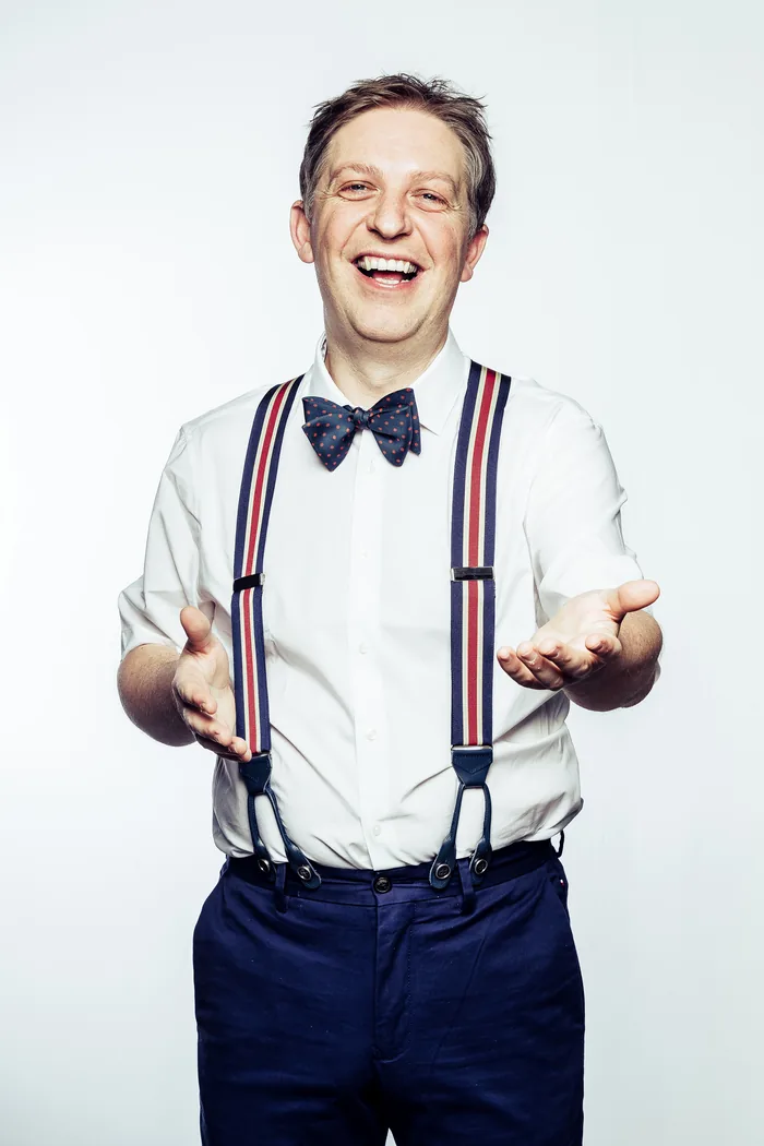 Manu Houdart, en chemise blanche, nœud papillon et bretelles, les bras ouverts.
